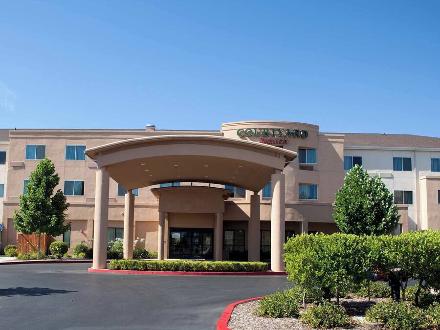 Un hotel Courtyard Marriott con entrada cubierta, varios pisos y un área ajardinada con árboles y arbustos.