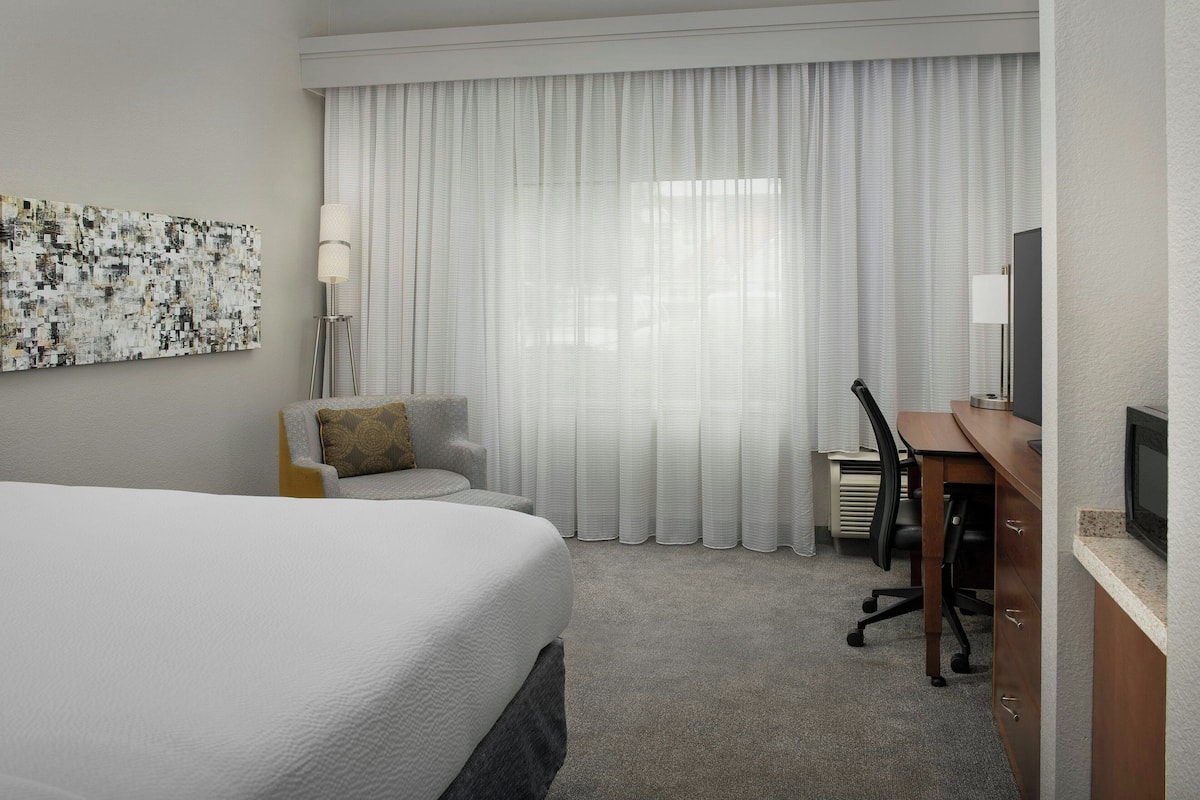 Una habitación de hotel con una cama grande, un escritorio con silla, un televisor y una ventana con cortinas traslúcidas.