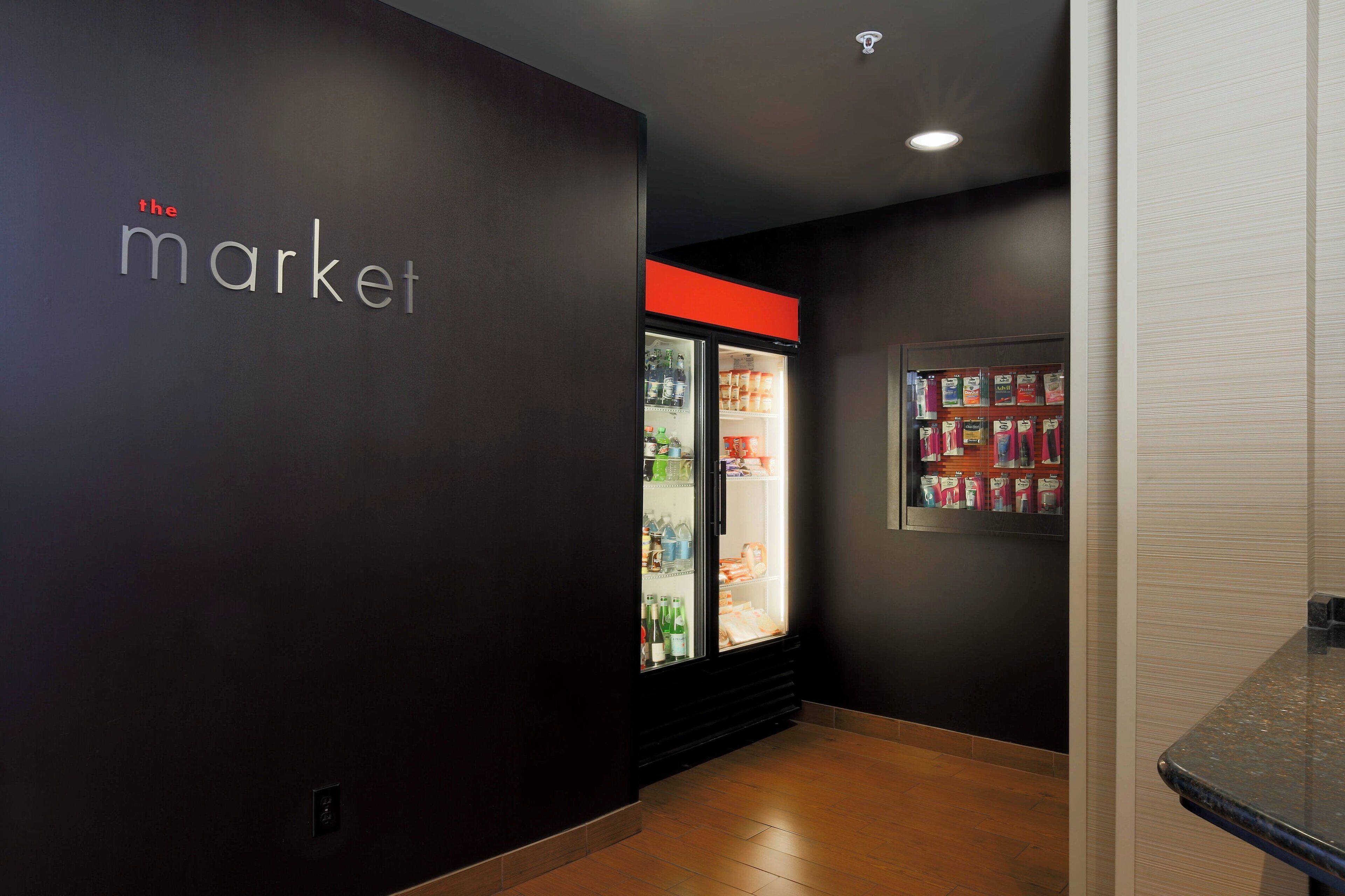 Um corredor com uma geladeira repleta de bebidas e lanches, uma parede com a inscrição 'the market' e um piso de azulejos.