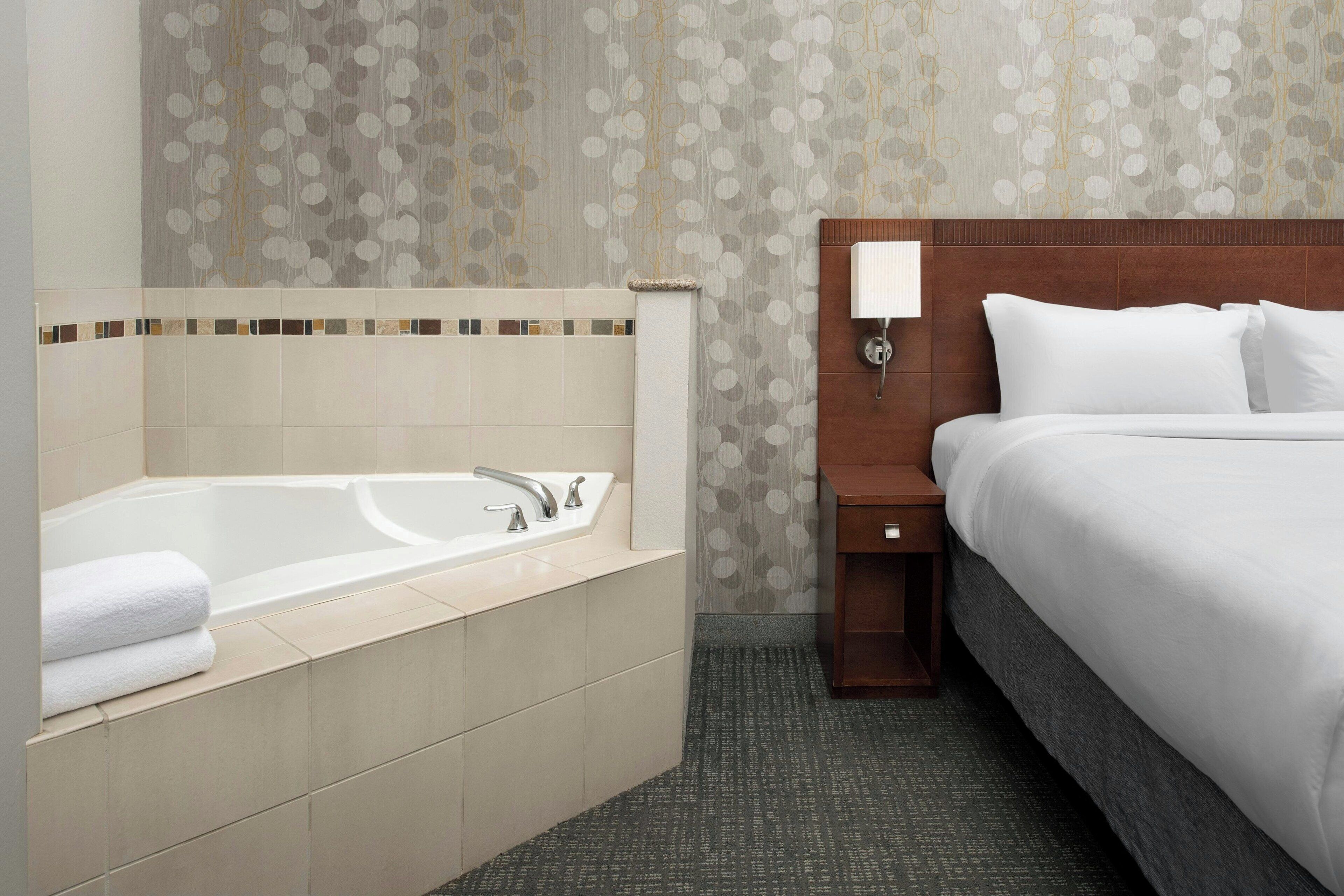 A hotel room with a bathtub, a bed, a nightstand, and a wall-mounted lamp.