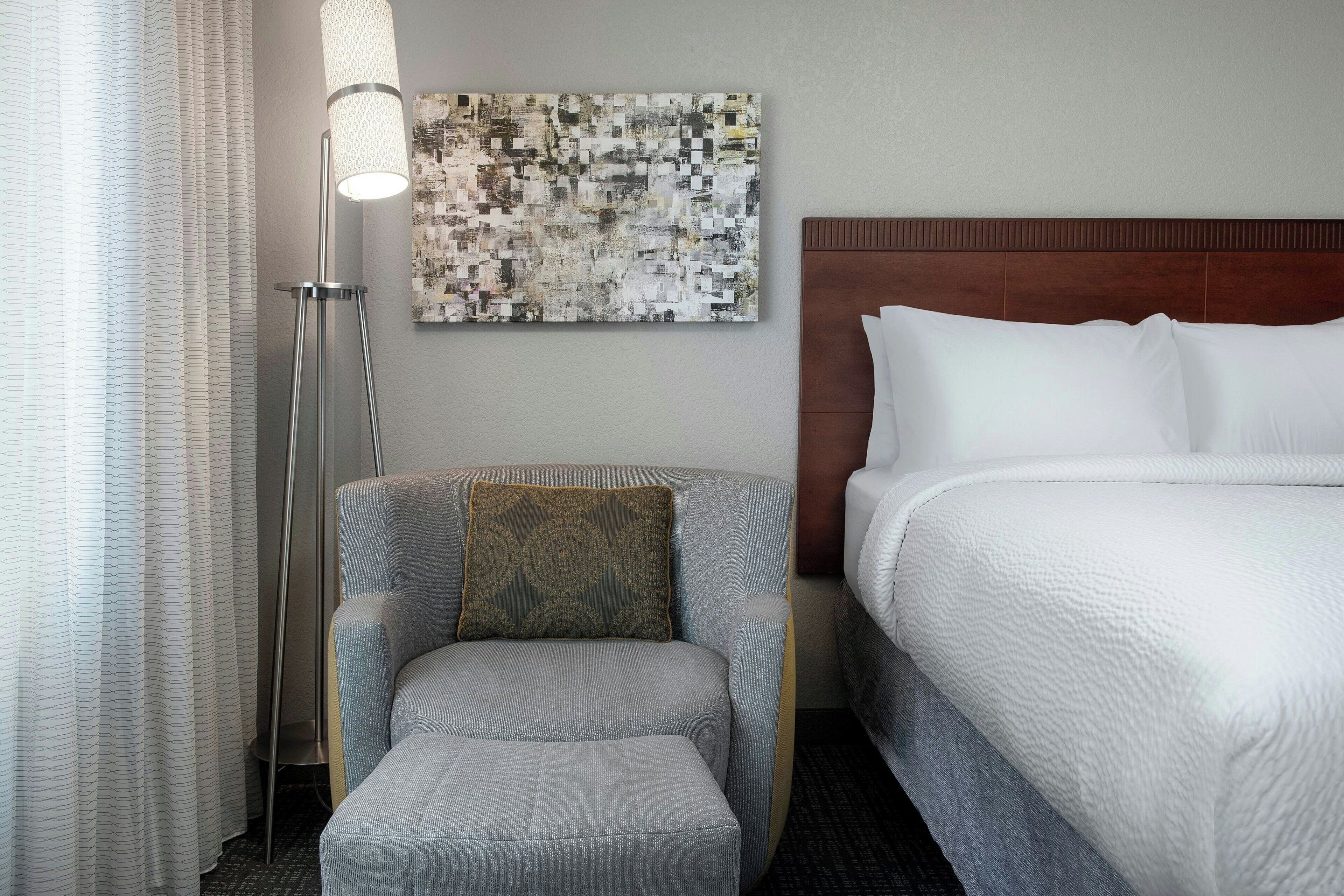 Una habitación de hotel con una cama, una silla, una lámpara de pie y una obra de arte en la pared.