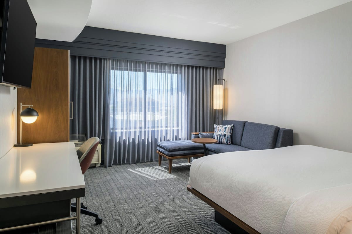 A modern hotel room with a large bed, a desk, a chair, and a sofa. There is a large window with curtains, a flat-screen TV mounted on the wall, and a lamp on the desk.