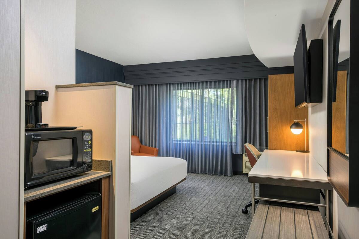 A hotel room with a bed, a desk, a microwave, a coffee machine, and a window with curtains.