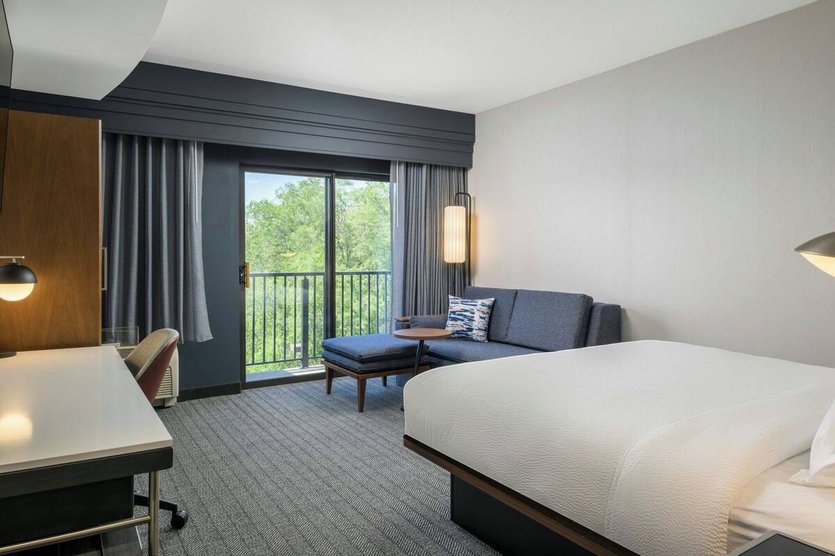 A hotel room with a large bed, a desk, a sofa, and a balcony with curtains.