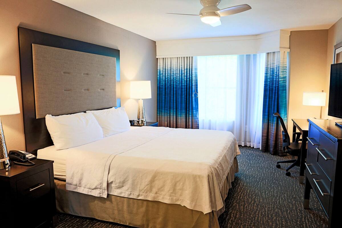 Ein Hotelzimmer mit einem großen Bett, zwei Nachttischen inklusive Lampen, einem Schreibtisch mit Stuhl und einem Deckenventilator.