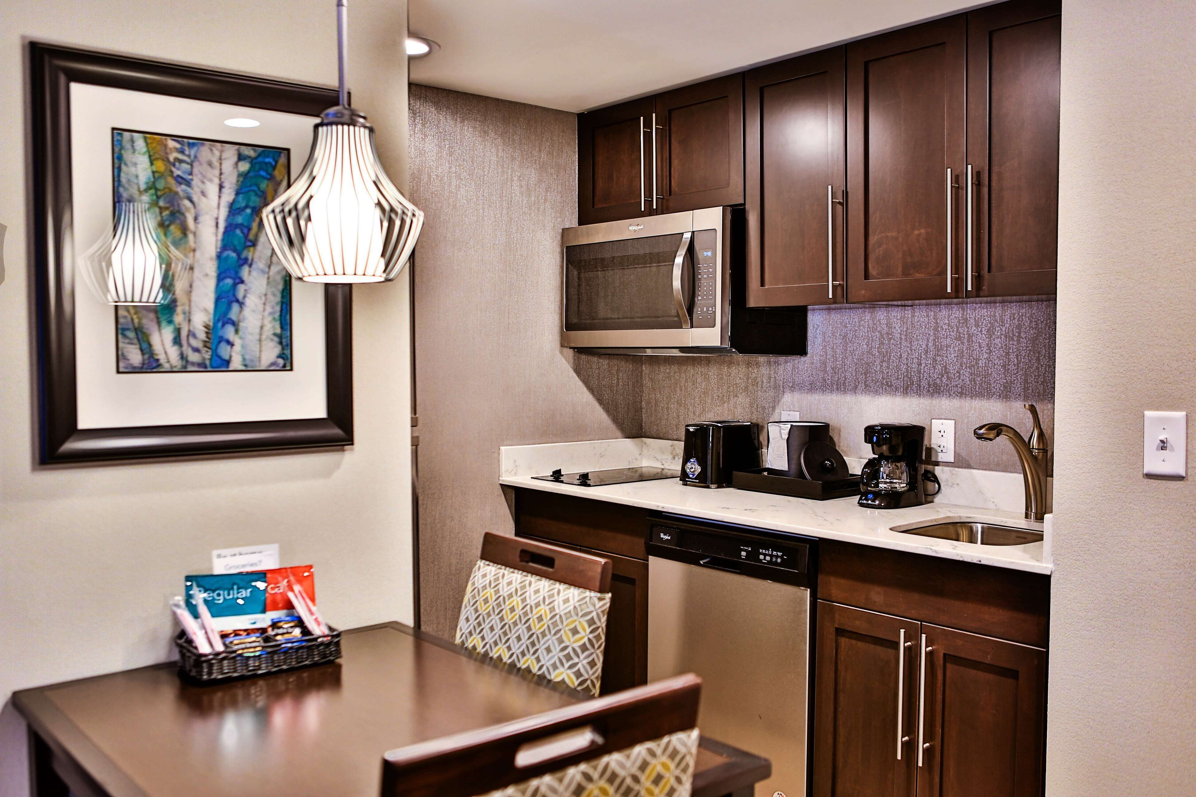 A compact kitchen with a microwave, coffee maker, and dishwasher. There is a framed artwork on the wall and a basket of toiletries on the table.