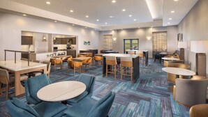 A hotel lobby with a mix of seating areas, including round tables and chairs, a kitchen area, and a bar.