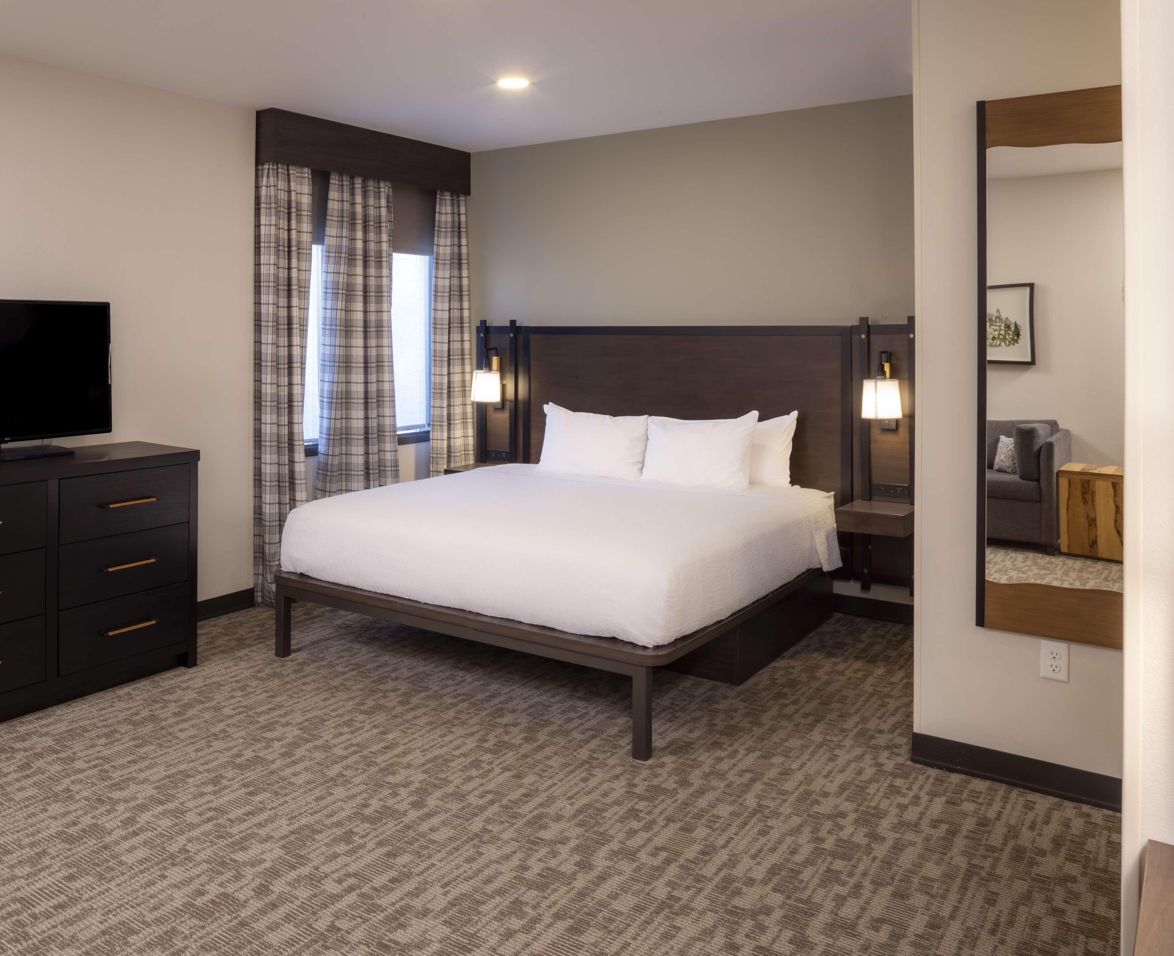 A hotel room with a large bed, a TV on a dresser, a nightstand with a lamp, and a mirror on the wall.