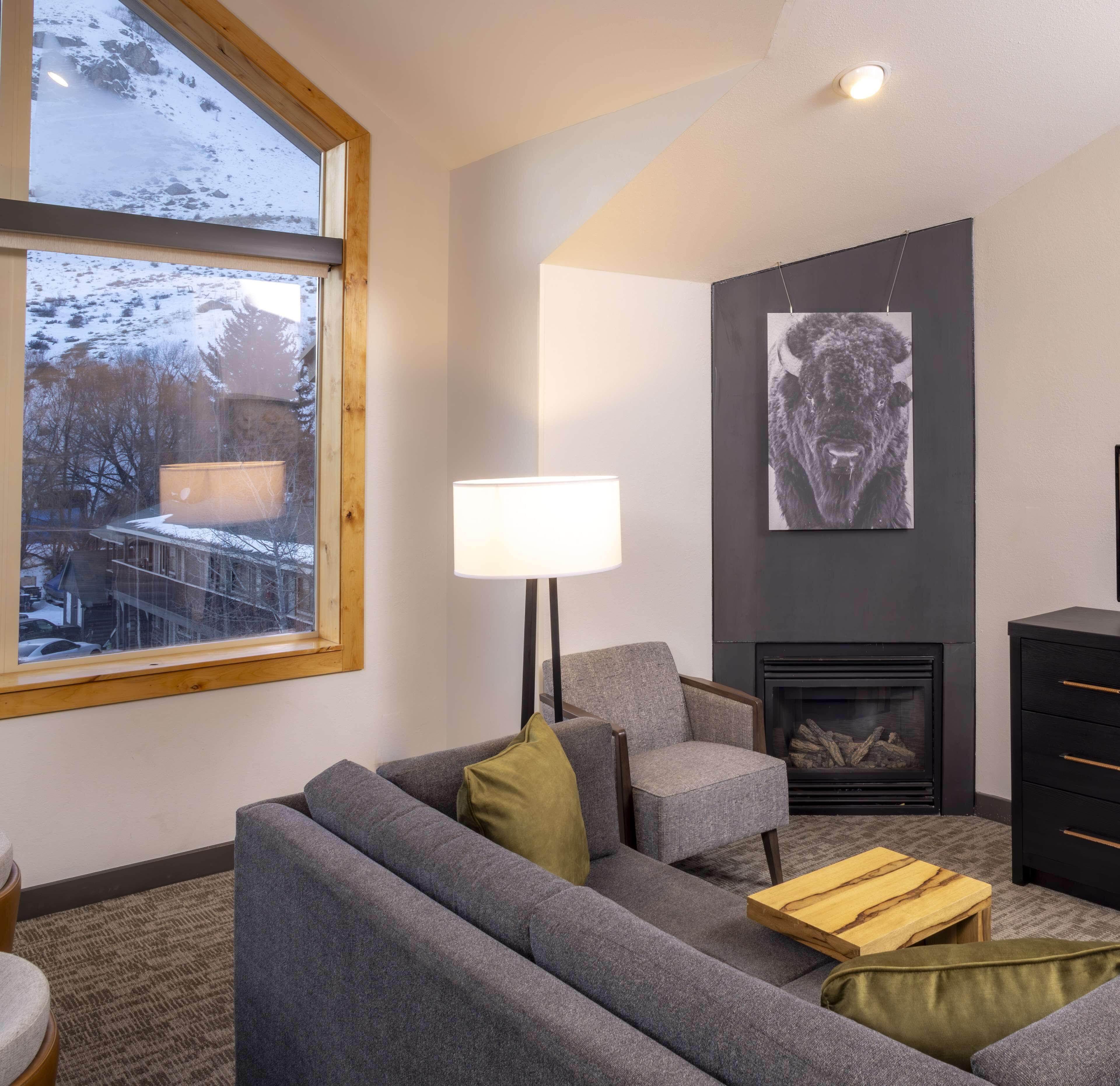 Ett modernt vardagsrum med en grå soffa, en lampa, en öppen spis och ett stort fönster med utsikt över ett snötäckt landskap.