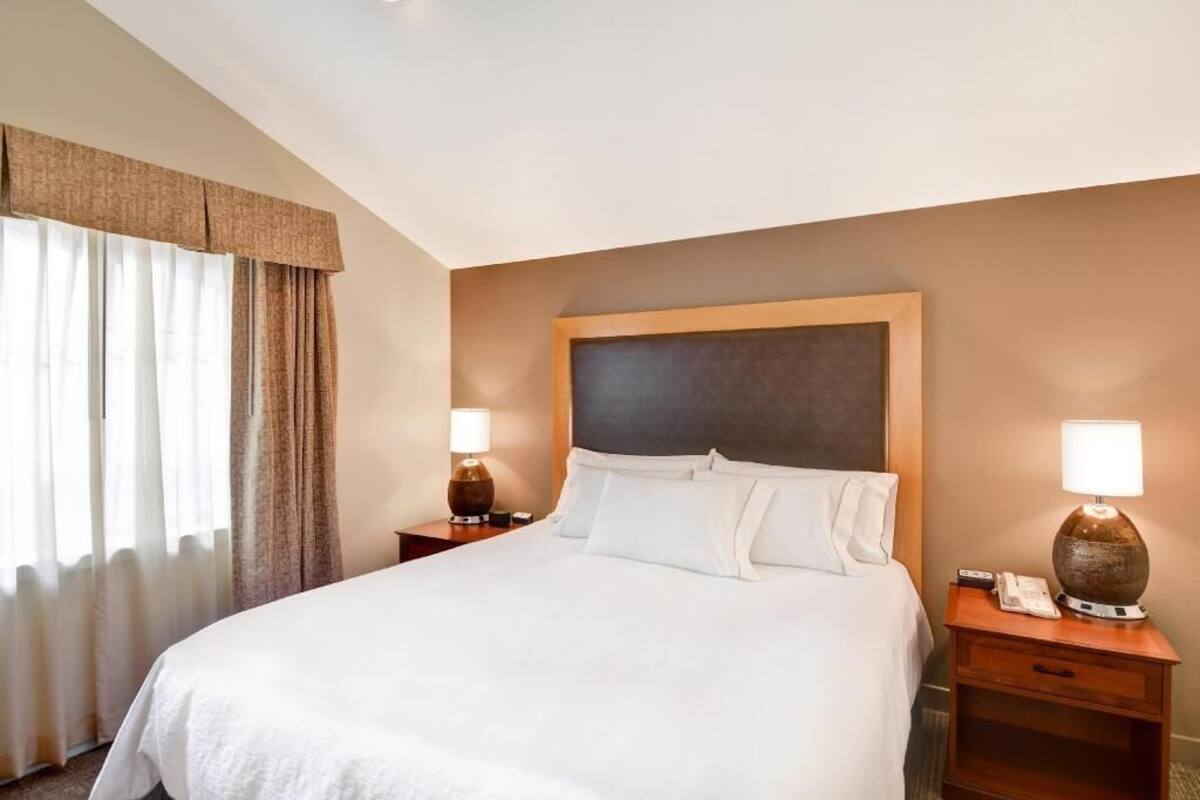 Una camera d'albergo con un letto grande, due comodini con lampade, una testiera e una finestra con tende.