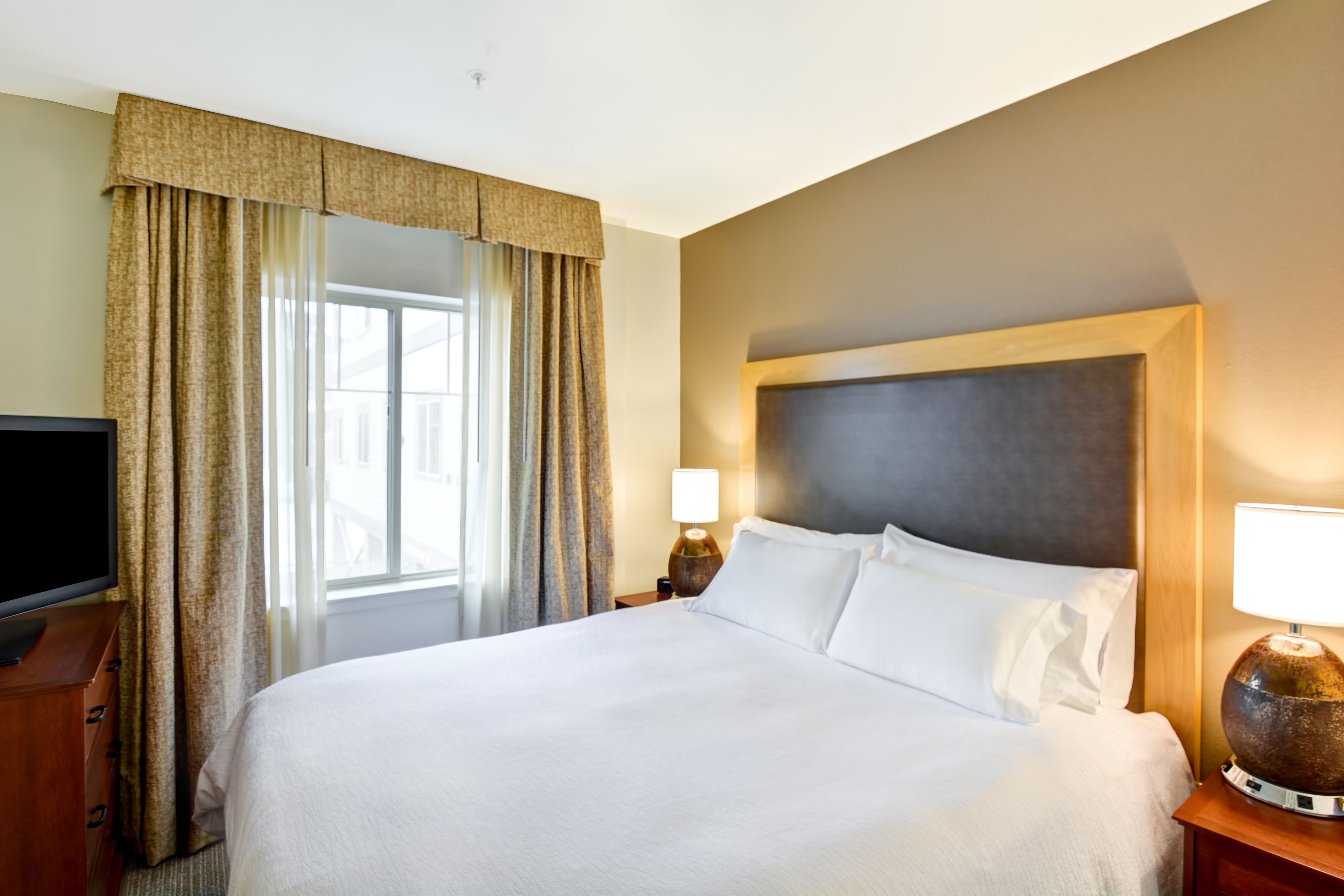 A hotel room with a large bed, a television on a wooden dresser, and a window with curtains.