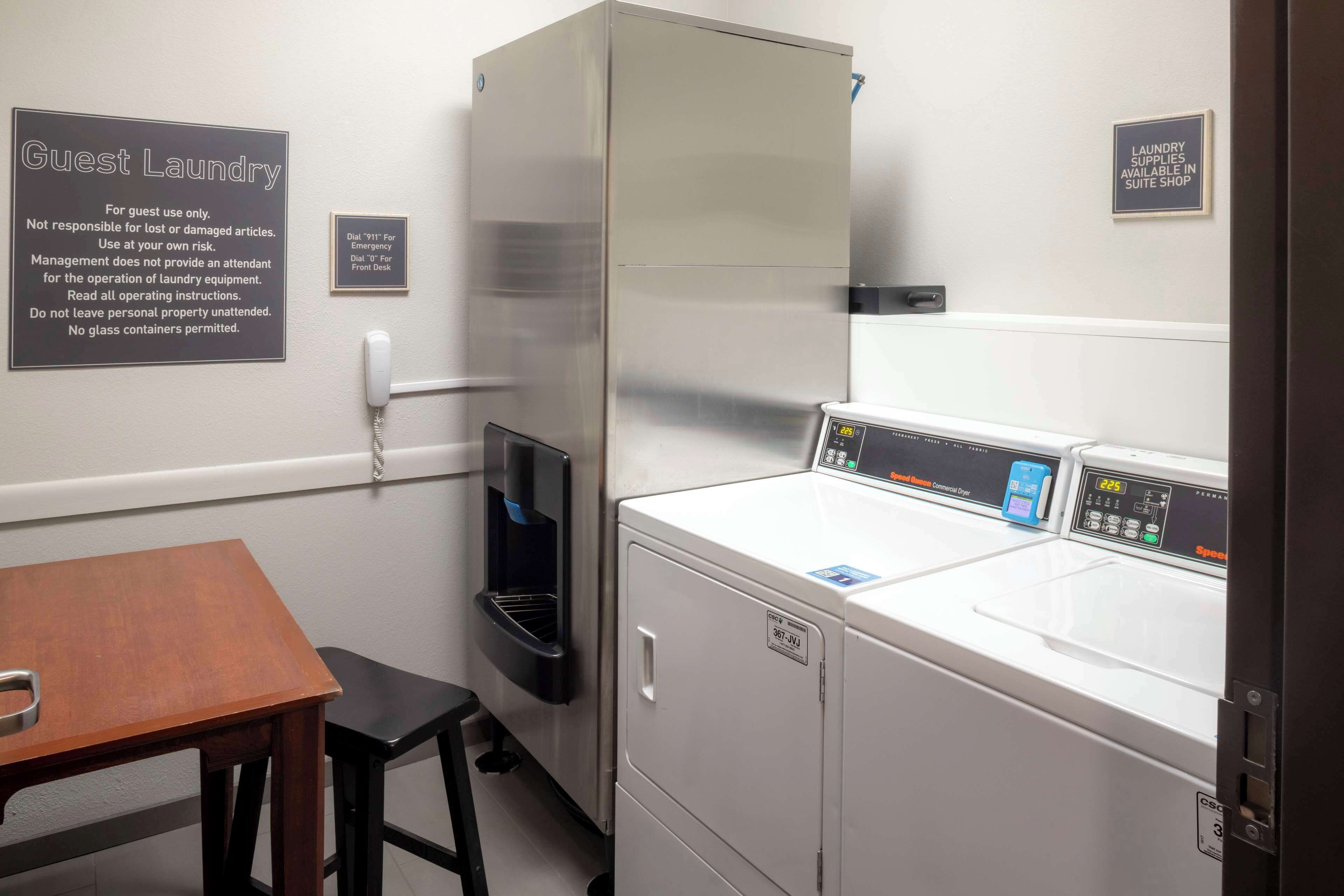 Een gemeenschappelijke wasruimte met een wasmachine, droger en een tafel met stoel. Bewegwijzering geeft instructies voor het gebruik van de wasfaciliteiten en een bericht over de beschikbaarheid van wasmiddel in de shop.