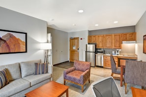 A modern living room with a sofa, armchair, and coffee table, a kitchen with wooden cabinets and appliances, and a framed artwork on the wall.