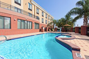 Una piscina con borde de ladrillo, un área de descanso con sillas y un edificio con múltiples ventanas y balcones.
