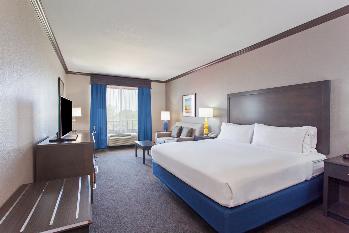 A hotel room with a large bed, a desk with a television, a sofa, and a window with curtains.