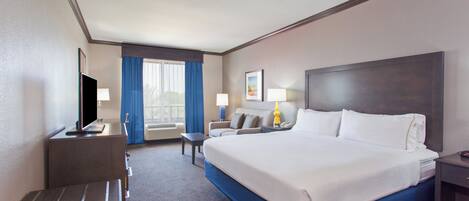 Habitación de hotel con una cama grande, un escritorio con televisor, un sofá y una ventana con cortinas.