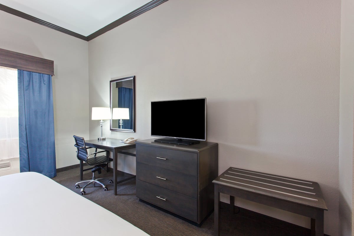 A hotel room with a bed, a desk with a computer, a TV, a chair, and a small table.