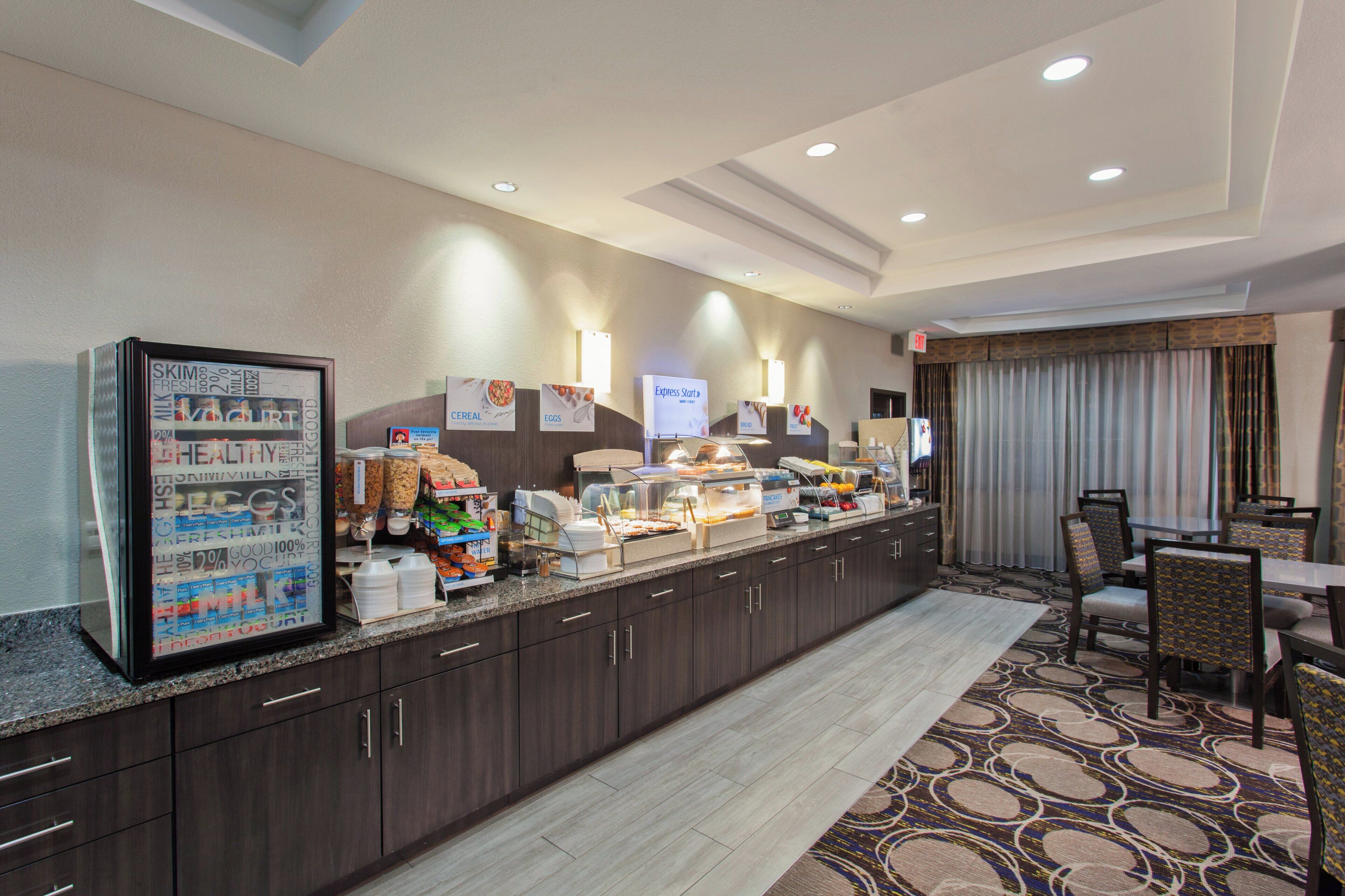 A hotel breakfast area with a self-serve station, seating, and a view of the room.