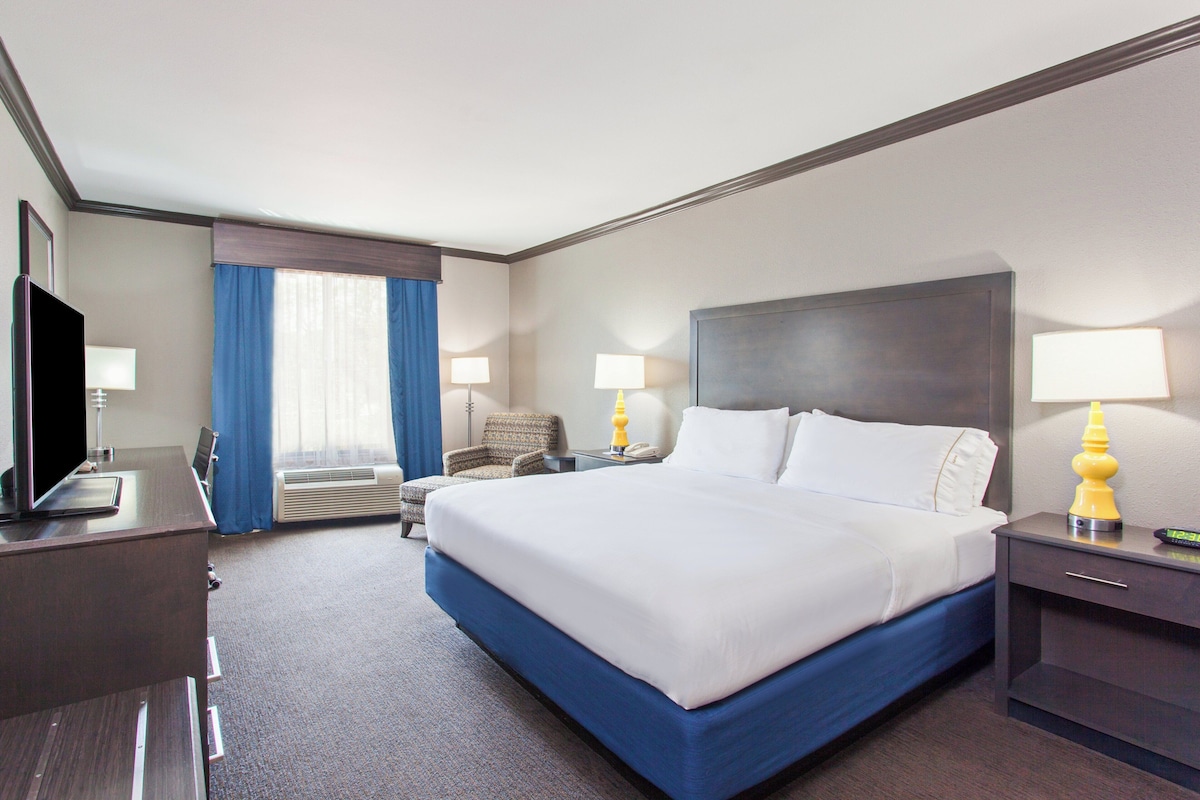A hotel room with a large bed, a desk with a flat-screen TV, a chair, two bedside lamps, and a nightstand with a clock.
