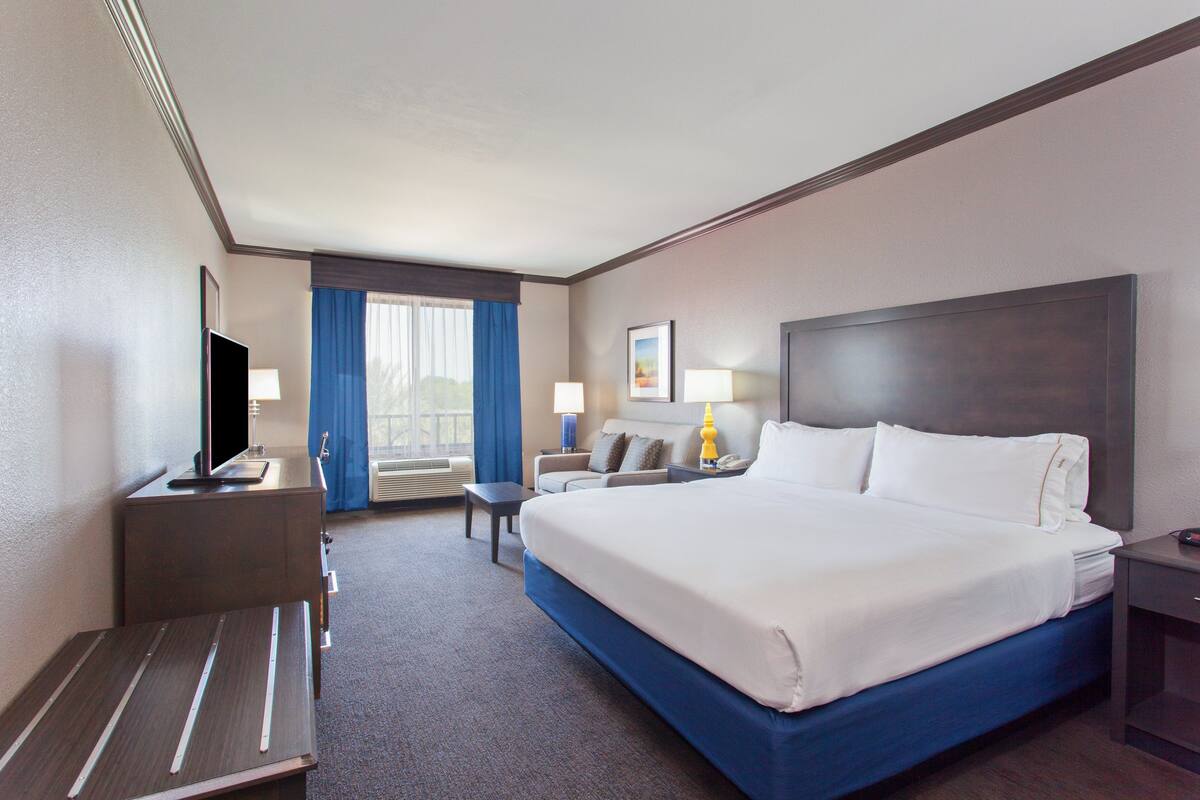 Une chambre d’hôtel avec un grand lit, un bureau avec une télévision, une chaise, un canapé et une fenêtre avec des rideaux.
