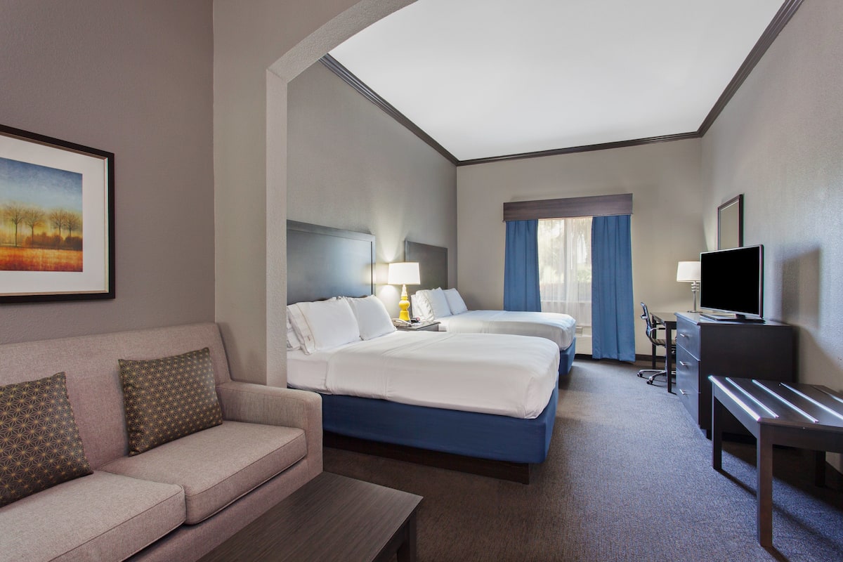 A hotel room with a sofa, two beds, a desk, a TV, and a painting on the wall.