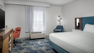 A hotel room with a bed, a desk, a chair, a TV, a window with curtains, an air conditioner, and a bedside lamp.