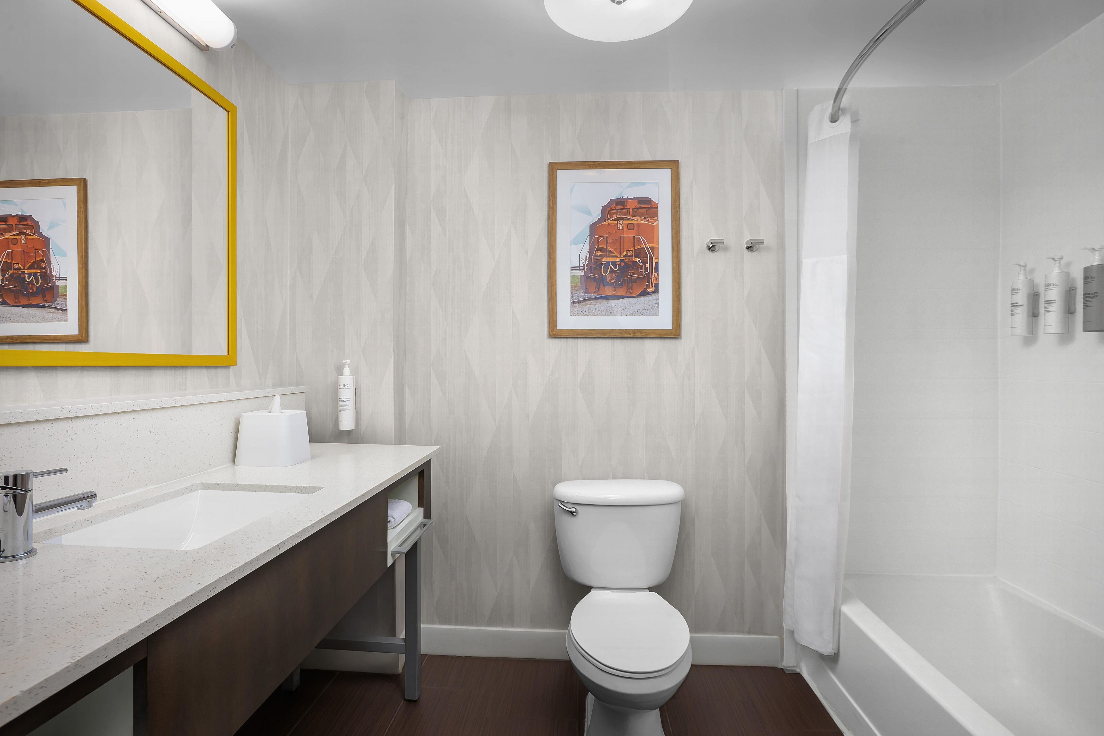 Une salle de bain moderne avec un lavabo, des toilettes et une douche, le tout de couleur blanche. Des tableaux encadrés ornent le mur, et un miroir à cadre jaune est également présent.