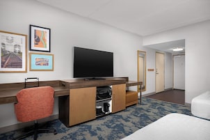 Habitación de hotel con televisor de pantalla plana, escritorio con silla, cama y baño visible a través de una puerta abierta.