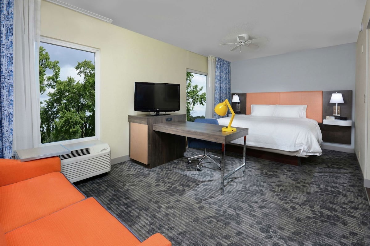 A hotel room with a bed, desk, chair, TV, and a view of trees through the window.