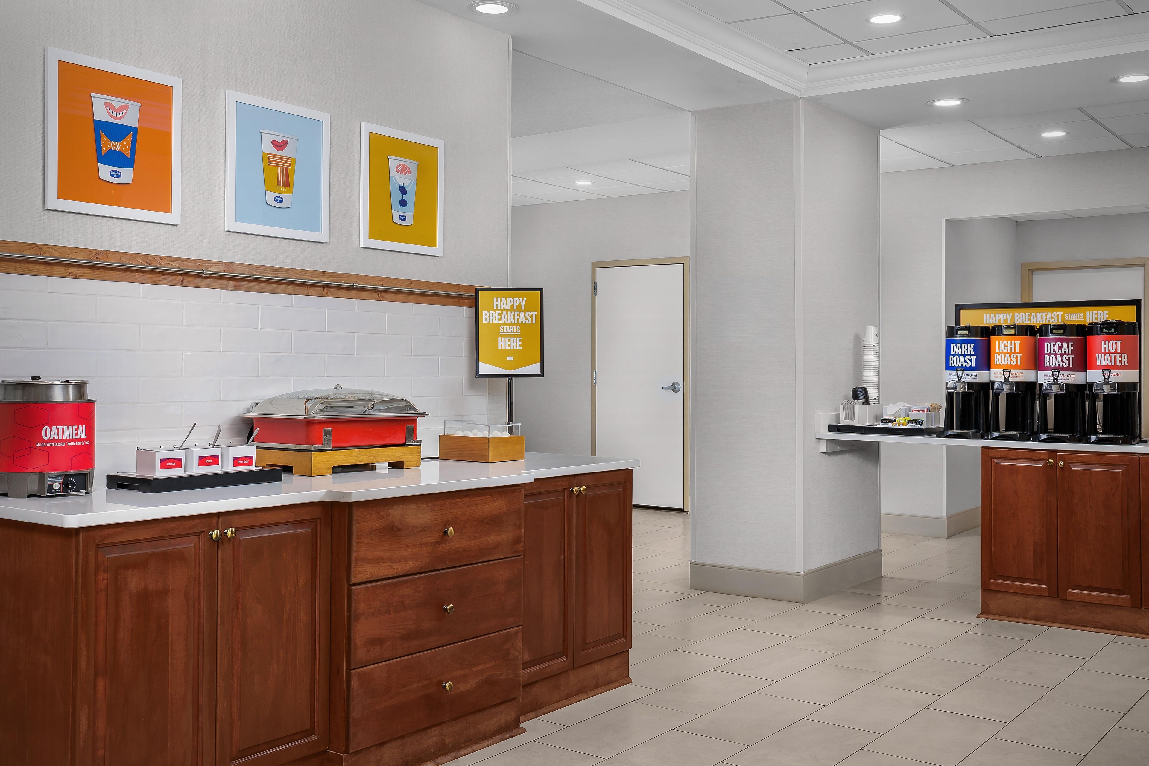 Un espace réservé au petit-déjeuner, avec un coin café, un panneau indiquant le début du service du petit-déjeuner, et un mur orné d’œuvres d’art encadrées.