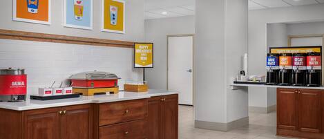 A breakfast area with a coffee station, a sign indicating breakfast starts here, and a wall with framed artwork.