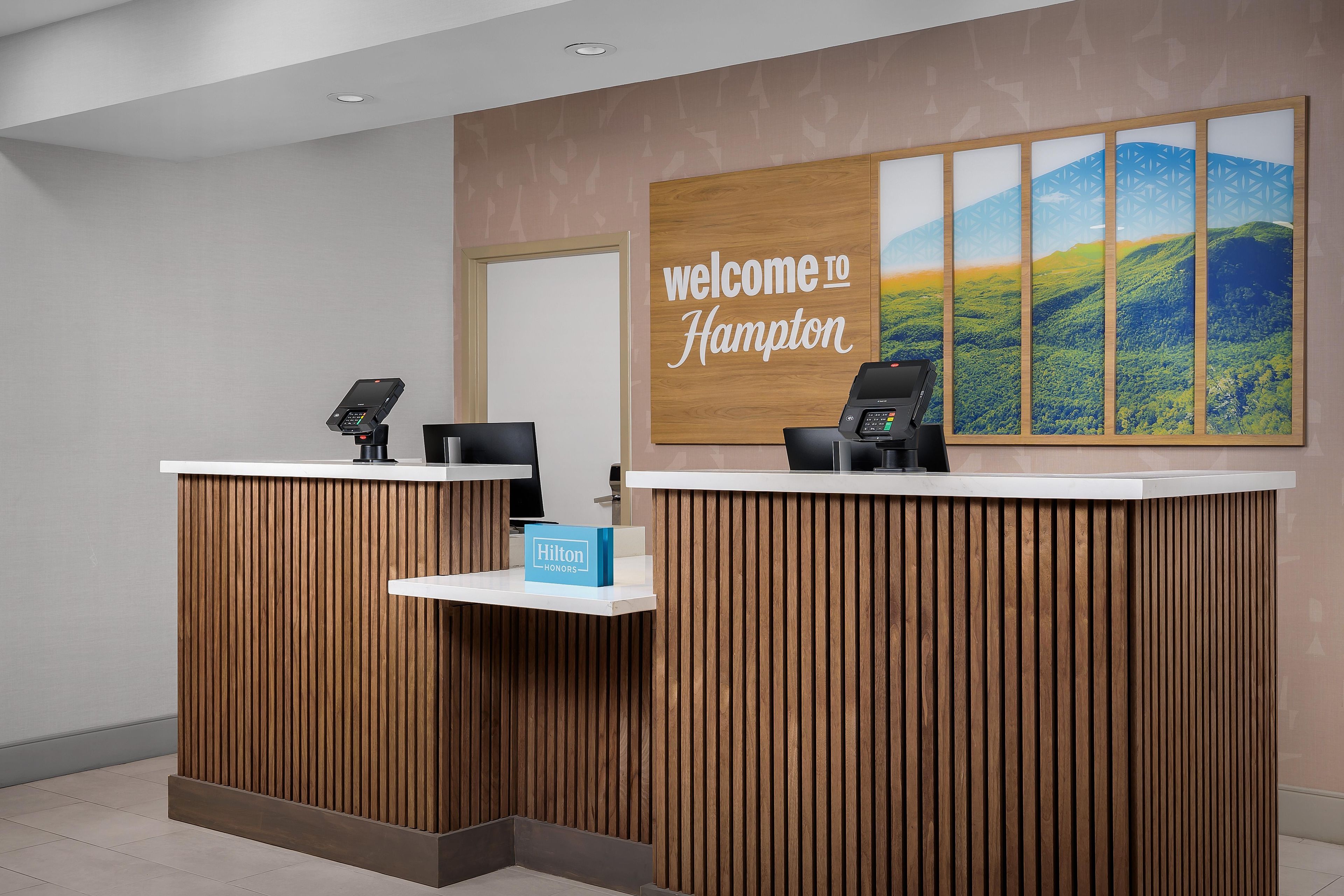 Un comptoir d’accueil avec une pancarte « Bienvenue à Hampton », une carte « Hilton Honors » et une vue panoramique sur les collines.