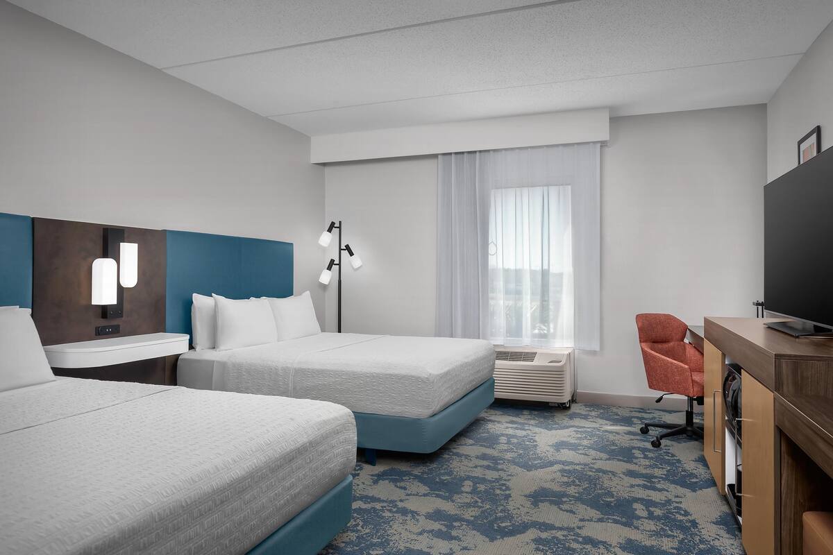 A hotel room with two beds, a desk with a television, a chair, a desk lamp, and a large window with curtains.