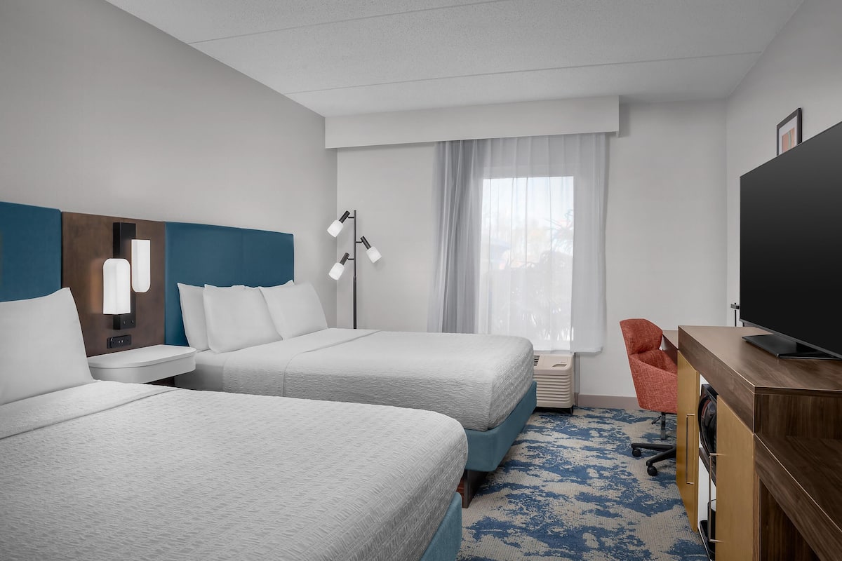 A hotel room with two beds, a desk with a TV, a chair, a lamp, and a window with curtains.