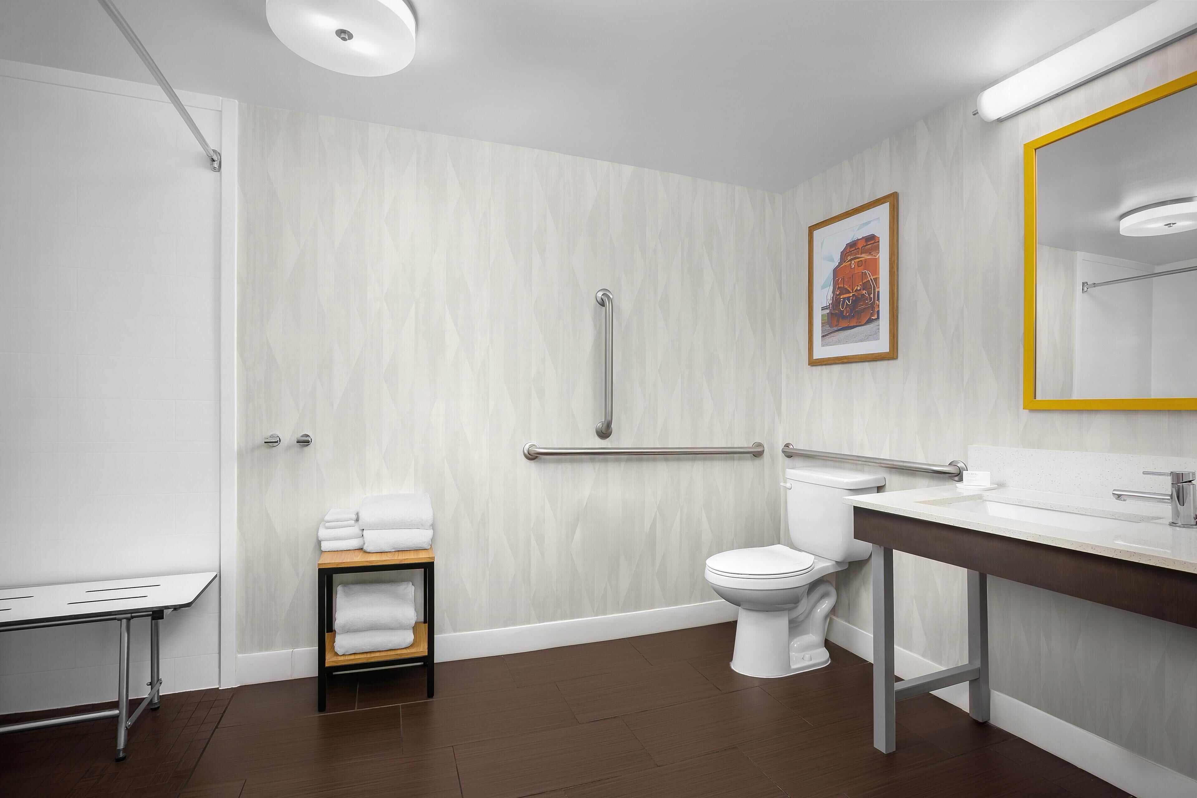 Un baño moderno con inodoro blanco, lavabo con espejo, ducha con barra de apoyo y un perchero para toallas.