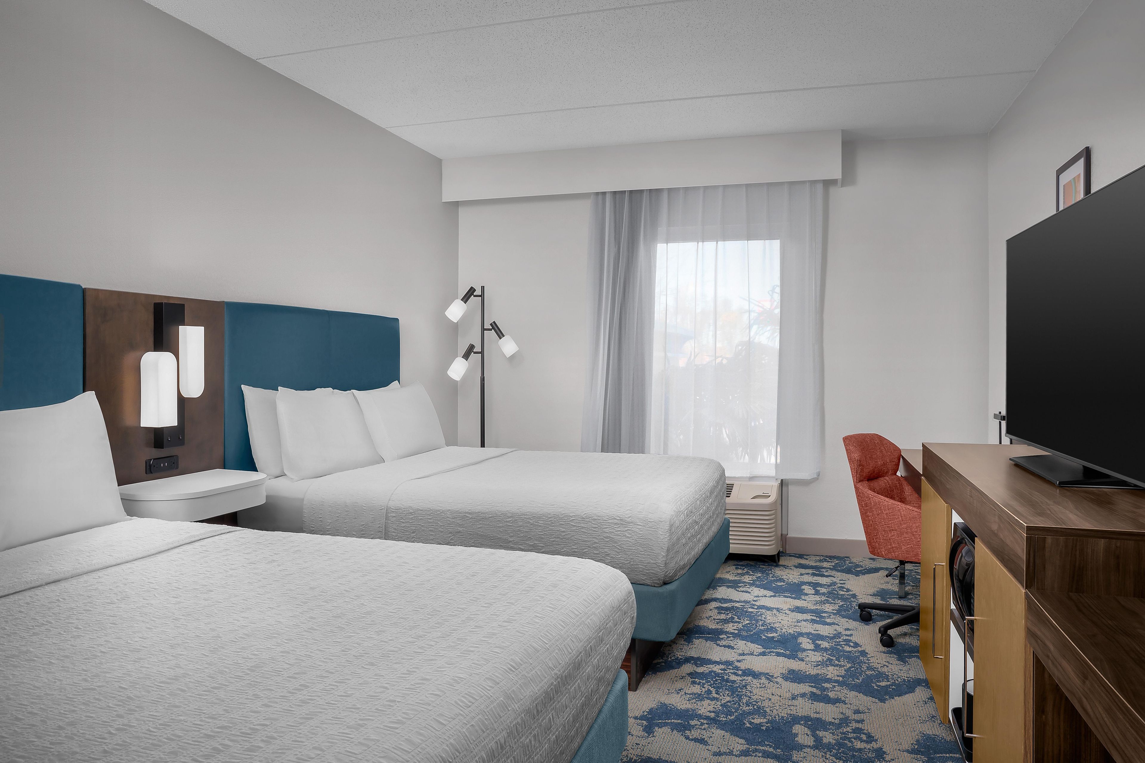 Ett hotellrum med två sängar, ett skrivbord med en platt-TV, en stol och ett fönster med gardiner.