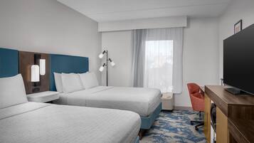 Ett hotellrum med två sängar, ett skrivbord med en platt-TV, en stol och ett fönster med gardiner.