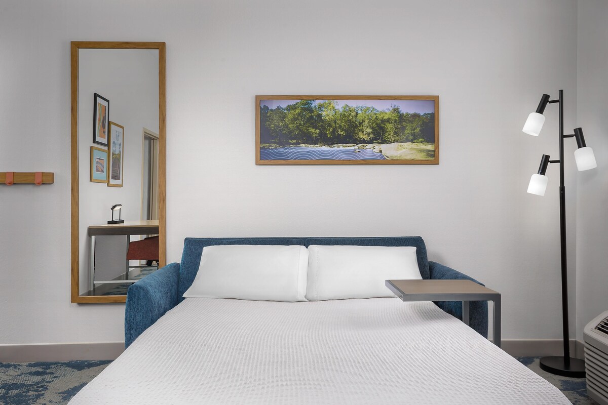 Hotellihuone, jossa on sänky, yöpöytä, peili, taulu seinällä ja lattiavalaisin.