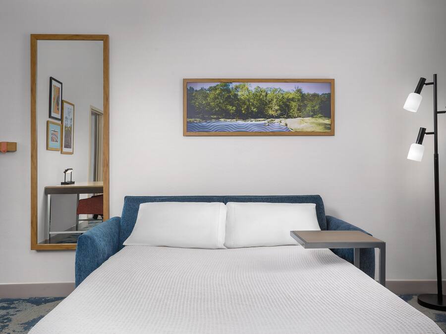 A hotel room with a bed, a bedside table, a mirror, a picture on the wall, and a floor lamp.
