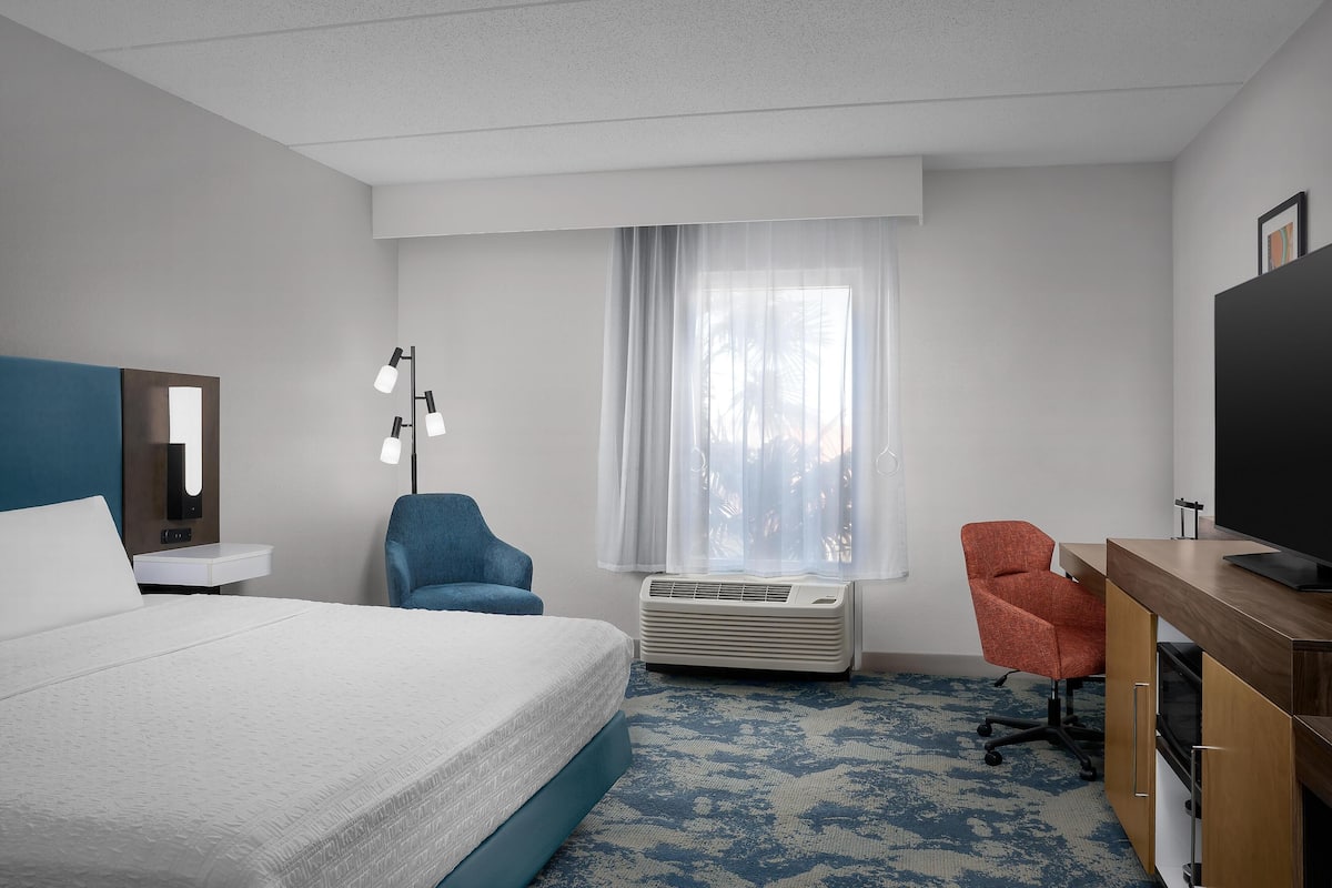 A hotel room with a bed, a desk with a flat-screen TV, a chair, a desk lamp, a window with sheer curtains, and an air conditioning unit.
