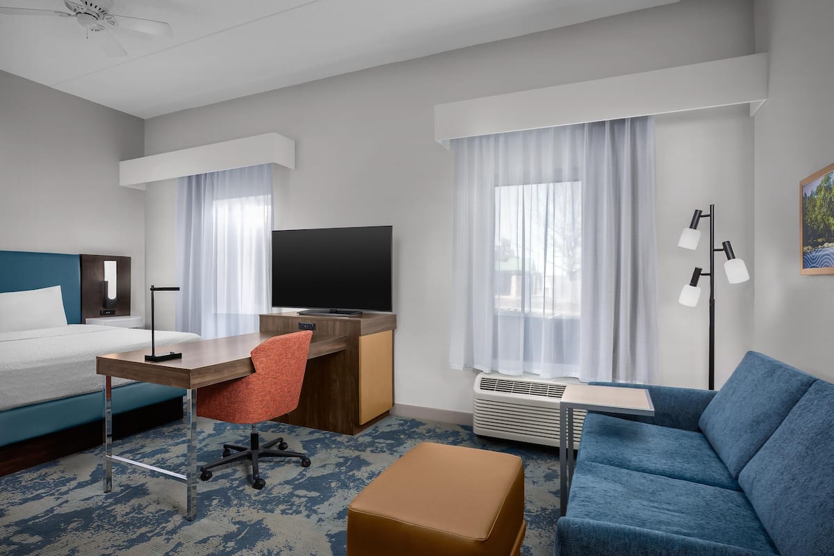 A hotel room with a bed, desk, chair, TV, sofa, and large windows with curtains.