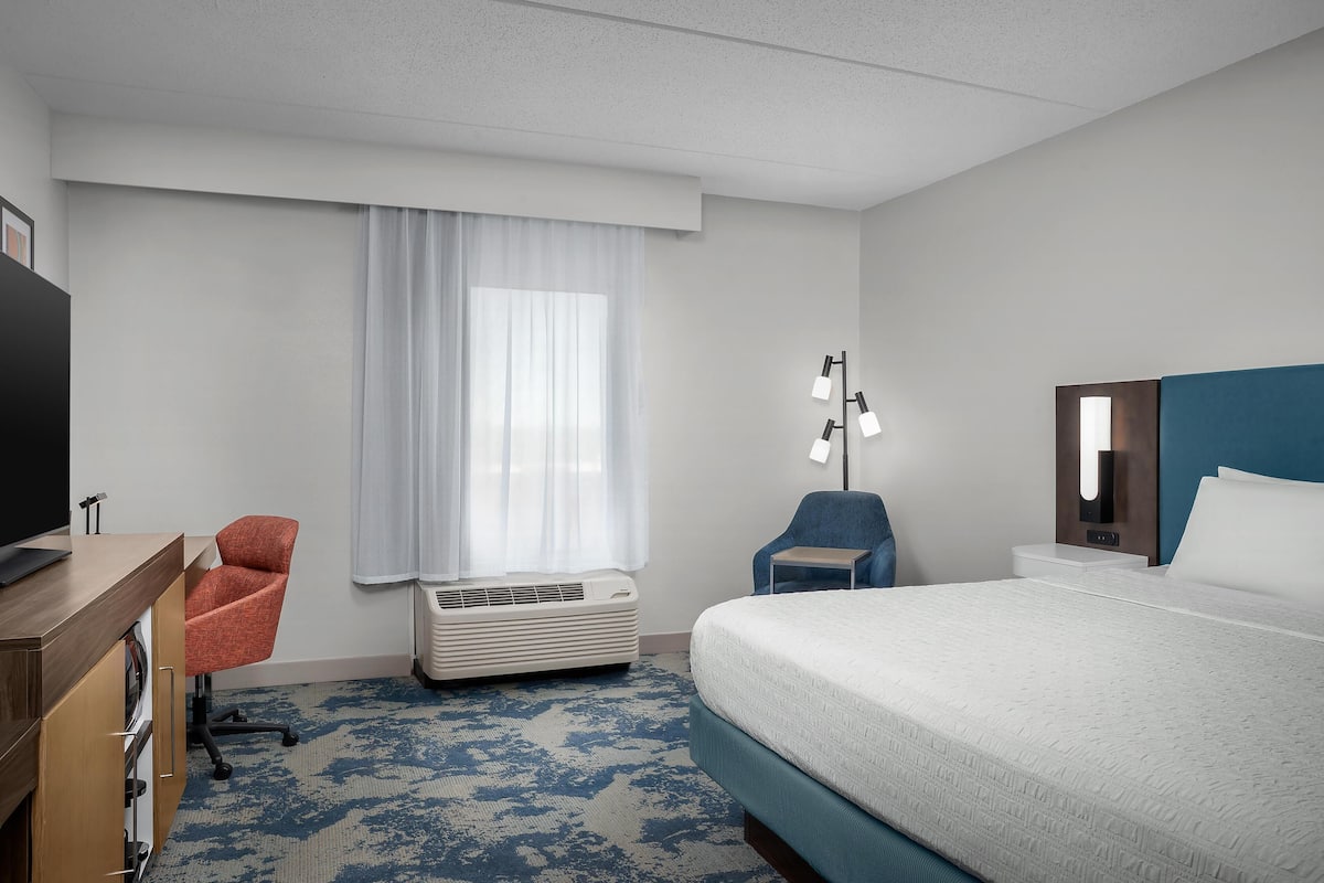 Une chambre d’hôtel comprenant un lit, un bureau avec une chaise, une télévision, un climatiseur, une lampe et une fenêtre avec des rideaux.