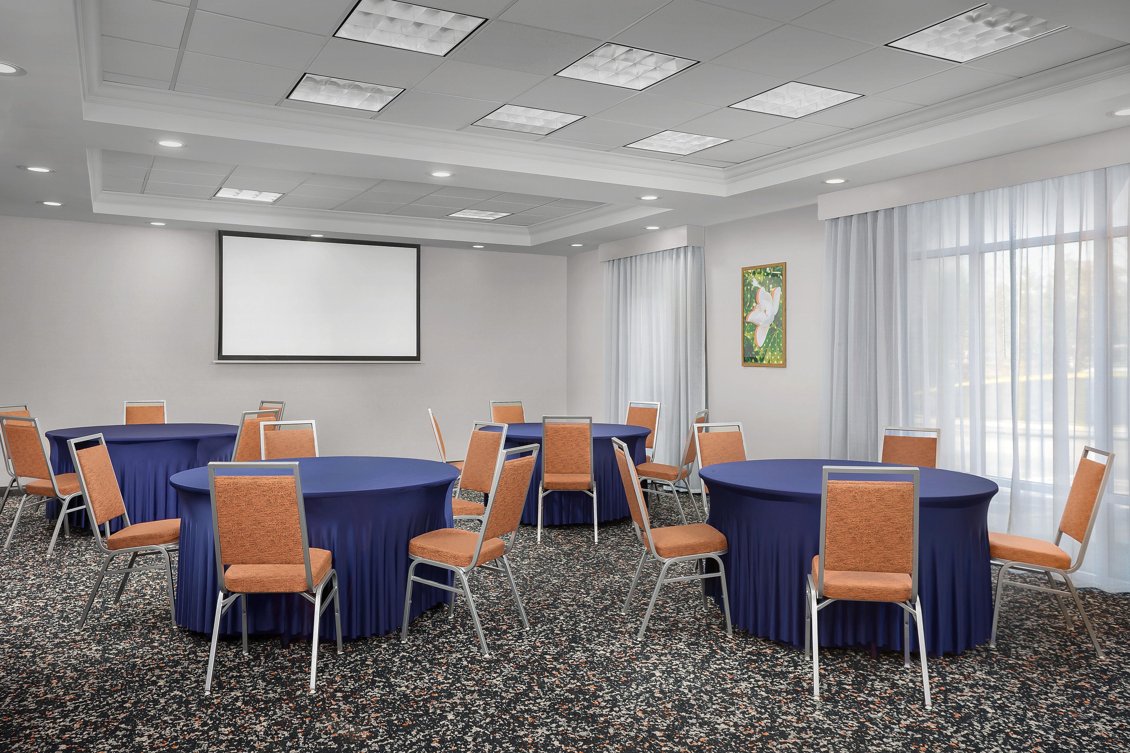 A conference room with round tables, chairs, a projector screen, and a large window with curtains.