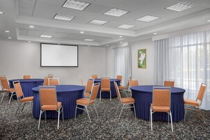 Un salón de conferencias con mesas redondas, sillas, una pantalla de proyector y un gran ventanal con cortinas.