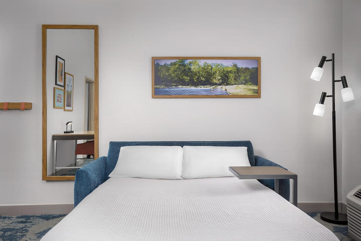 A hotel room with a bed, a desk, a mirror, a picture on the wall, and a floor lamp.