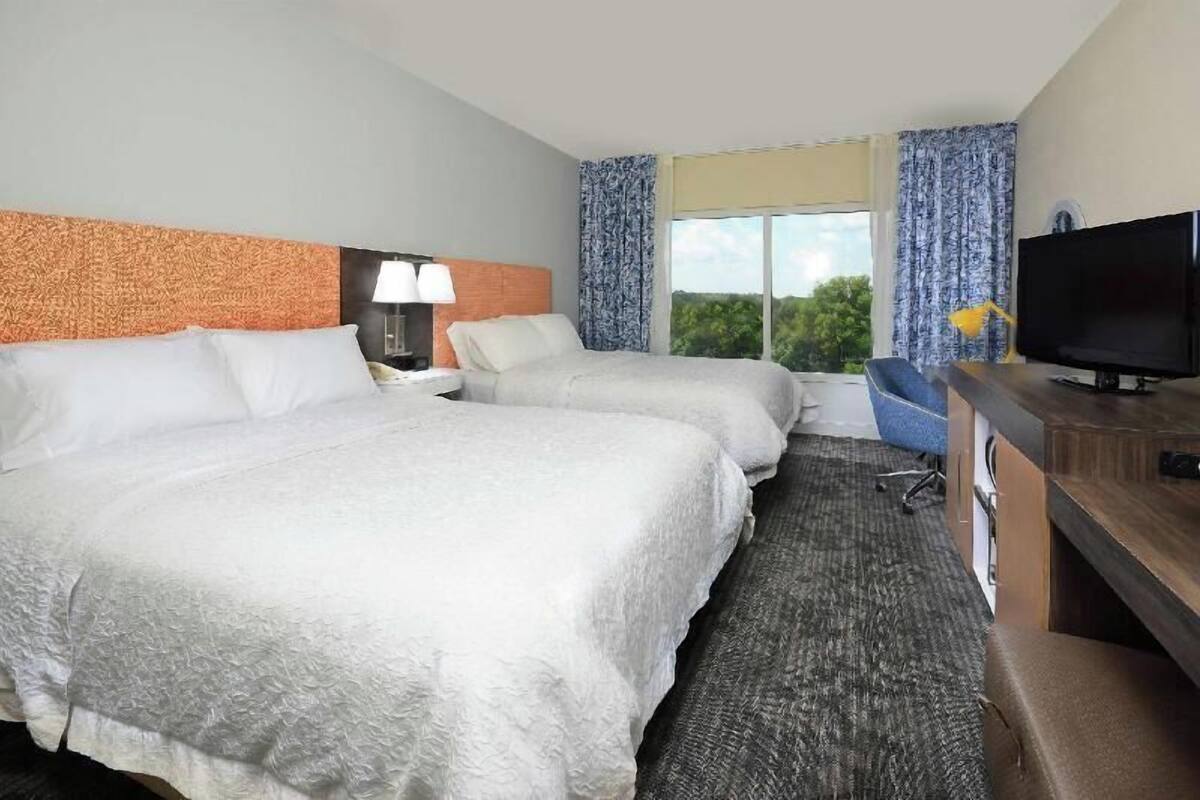 A hotel room with two beds, a television, a desk, and a view of the outdoors.