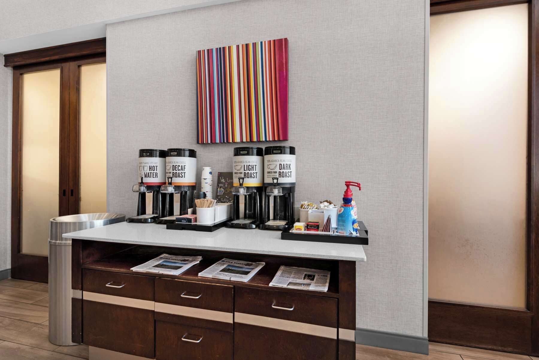 Une chambre d’hôtel équipée d’un coin café avec quatre distributeurs de café étiquetés « EAU CHAUDE », « CAFÉ DÉCAFÉINÉ », « CAFÉ LÉGER » et « CAFÉ CORSÉ ». On y trouve également des journaux sur le comptoir, une poubelle et une bouteille de gel hydroalcoolique.