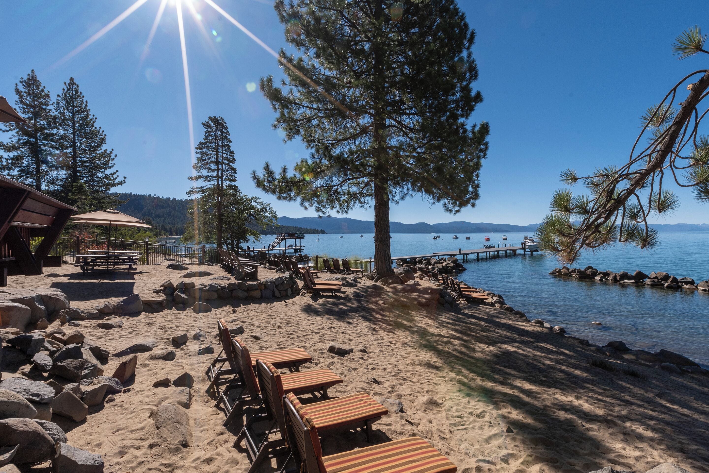 En strand med träsolstolar, ett trädäck och utsikt över sjön och bergen.