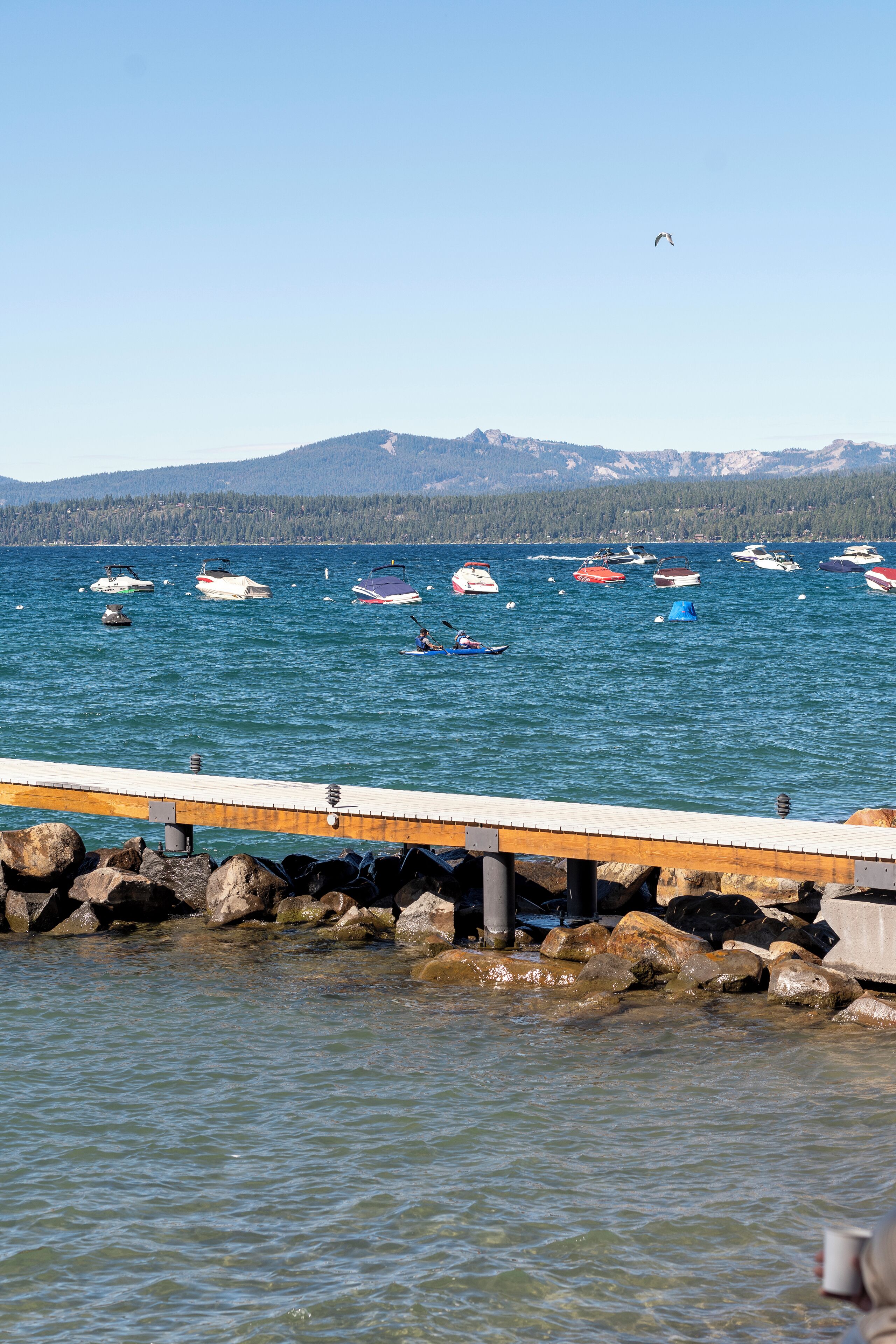 A pier with boats on a calm lake, surrounded by rocky shores and distant mountains.