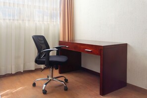 Un escritorio de madera con una silla de oficina negra, un cajón y una ventana con cortinas traslúcidas.